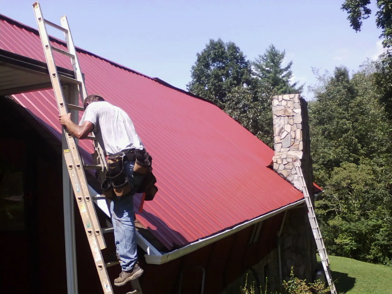 Roofer on a ladder beside a red metal roof and stone chimney — Soffit & Fascia Repair by Roofing Brothers in Brock